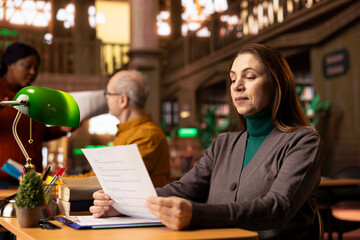 Elderly woman reading and studying in a campus library with materials, immersed in learning and surrounded by academic books. Senior female learner in higher education environments.