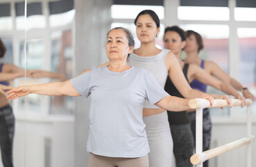 Women of different ages exercise near the ballet barre at group training session in the batman...