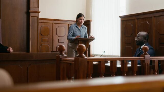 Young woman in light blue shirt testifying in court, while lawyers, judge, and defendant listening to her