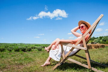 Young beautiful sexy woman in straw hat and sunglasses sitting on a sun lounger on a meadow on a...