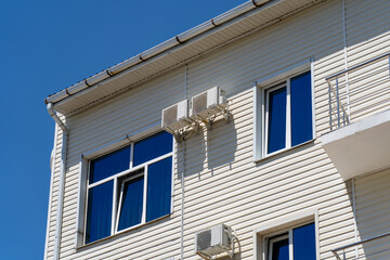 Bright Day View of a Modern Building Exterior, Showcasing Light Siding, Reflective Blue Windows, and Outdoor Air Conditioning Units Against a Clear Sky for Architectural Use.
