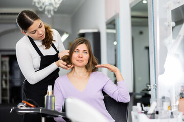Young woman hairdresser examining the hair of a female client in a chair in a barber shop,...