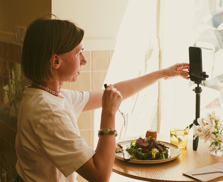 Young woman eating salad with meat during lunch break in cafe and live streaming on social media.  Soft focus.  Close-Up.