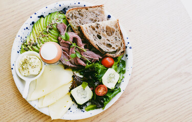 Salad, a piece of red meat, butter, a few slices of cheese, bread and a boiled egg lie on a plate standing on the table. Healthy food. Healthy lifestyle. Close-up. Soft focus