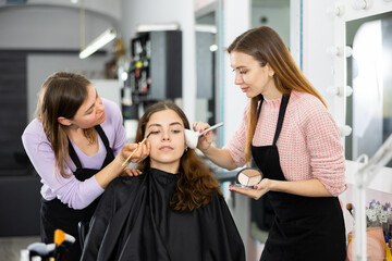 Focused professional women makeup artists applying shadows and blush with brushes on face to young...