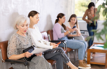 Elderly woman in casual clothes reads text of document while sitting on chair in reception waiting...
