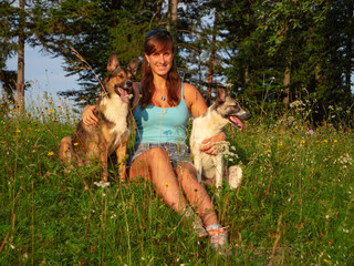 PORTRAIT: Smiling woman sits in a sunlit meadow by the forest, embracing her two adopted dogs....
