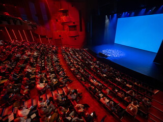 Theater auditorium interior with red seats and dramatic blue stage lighting showing audience and performance space