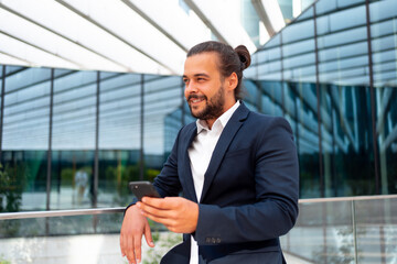 Businessman standing outdoor use smartphone looking away and smile. Handsome hispanic male business...