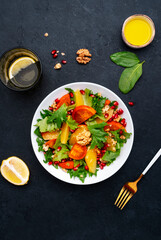 Autumn vegan persimmon salad with pomegranate seeds, walnuts, orange slices and lettuce, black background, top view