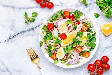 Gourmet seafood salad with squid, juicy orange, red tomatoes, lamb lettuce on marble table, top view
