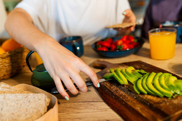 Woman using a butter knife to prepare sliced avocado on a wooden cutting board during breakfast at...
