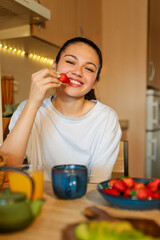 Happy young woman eating fresh strawberries for breakfast, enjoying a healthy meal in her kitchen