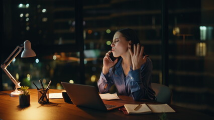 Overtime manager talking cellphone looking laptop at night office. Worried woman