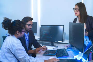 Diverse team of AI developers collaborating in a modern office. Indian male leading a discussion with Asian and Hispanic female colleagues on a machine learning project.
