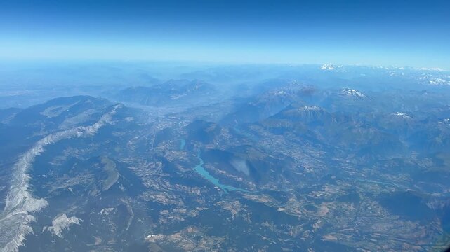 Sweeping aerial view of the European Alps from an aircraft at high altitude, revealing rugged mountain ranges, deep valleys, snow-covered peaks, and the expansive alpine landscape stretching toward th