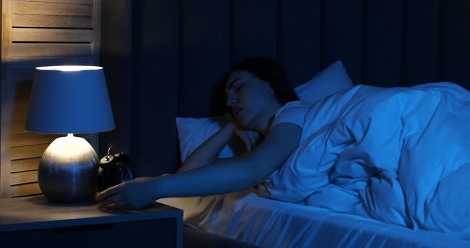 Frustrated woman with alarm clock suffering from insomnia in bed at home, camera moving down