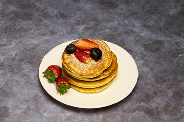 A plate of delicious pancakes in a tower with powdered sugar, decorated with blueberries and fresh berries (strawberries) for a wonderful breakfast.