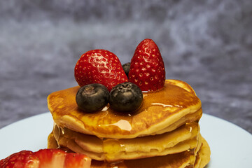 A plate of delicious pancakes in a tower with powdered sugar, decorated with blueberries and fresh berries (strawberries) drizzled with delicious maple syrup for a wonderful breakfast.