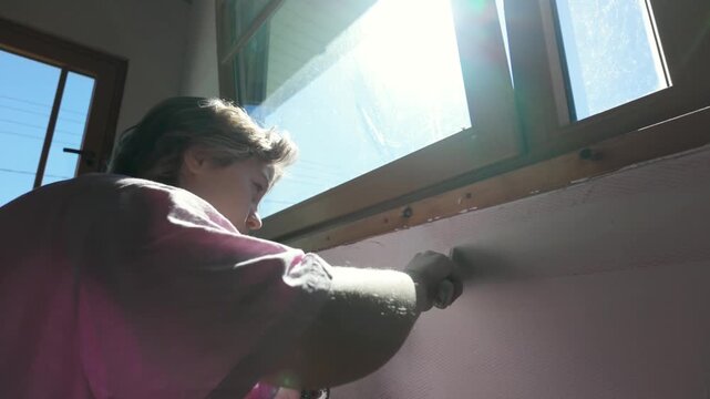 Teenage girl spreading putty across wall surface. Filling cracks with spackle, preparing walls for painting. Helping parents with home renovation. Slow motion sunny bright indoor footage.