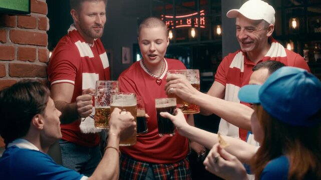 Two groups of happy young football fans in red and blue sportswear clinking with glasses of beer in pub while meeting each other and discussing latest match broadcast
