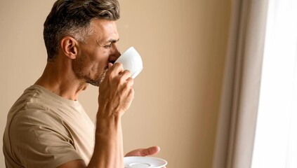 a man in a neutral room holding a cup of coffee, enjoying his coffee in the morning near window. He takes a moment of relaxation