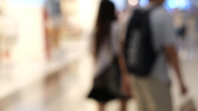 A blurry image of a man and woman walking in a mall with a backpack. The man is wearing a blue shirt and the woman has long hair. The backpack is black and has the word "adidas" on it
