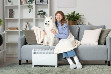 Young woman with Samoyed dog and plaid on sofa warming near radiator at home