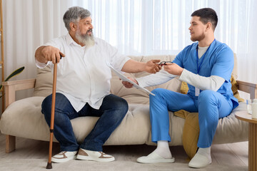 Obraz premium Young caregiver taking newspaper with eyeglasses to senior man at home