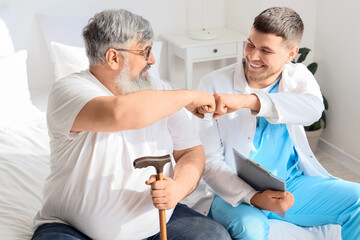 Young caregiver with senior man bumping fists in bedroom