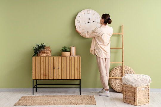 Young woman hanging clock on green wall near chest of drawers in room