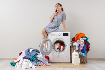 Young woman talking by phone while sitting on washing machine with pile of laundry against grey background