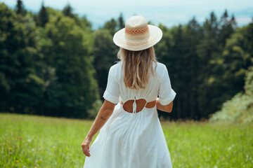 Rear view of a woman with long blonde hair in a white dress walking in a summer field with a straw...