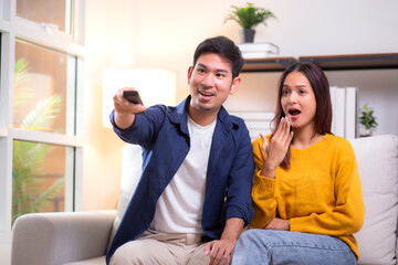 Obraz premium Young asian couple sitting on sofa in living room at home, reacting with surprise while watching television, expression emotional response during a dramatic scene, leisure together in cozy.