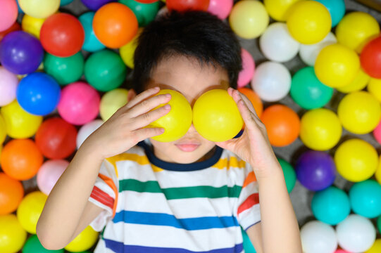 Playful asian boy lying in colorful ball pit covering his eyes with two yellow balls, reative and imaginative child having fun, indoor playground, showing sense of humor and joyful playtime