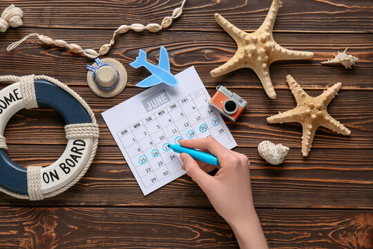 Female hand with marker, calendar and beach decor on wooden background. Summer vacation concept