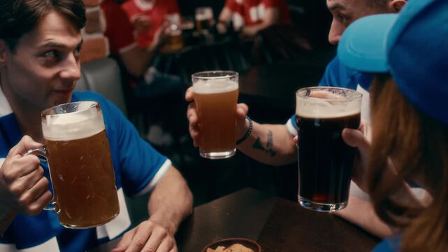 Group of three young football fans discussing latest match broadcast and having beer while sitting by table in bar and clinking with foaming beverage