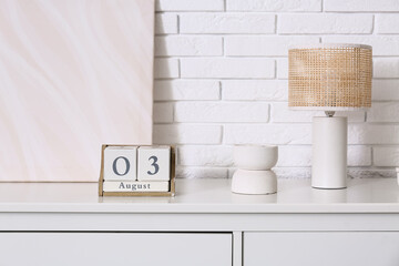 Cube calendar with date 3 AUGUST, lamp and painting on chest of drawers near white brick wall
