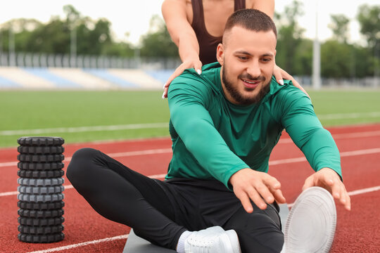 Sporty man with personal trainer stretching at stadium