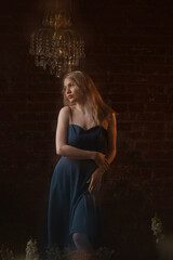 Young Caucasian woman in blue dress posing with raised arms, looking away. Front view. Vintage room with brick wall, crystal chandelier, flowers. Concepts: elegance, beauty, nostalgia, romance.