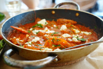Shakshuka dish simmering in a pan with fresh ingredients like tomatoes, peppers, and crumbled cheese. The vibrant tomato sauce is garnished with herbs and spices, ready for serving.