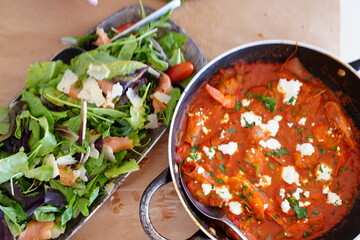 Vibrant mixed salad with greens, shaved cheese, and tomatoes beside a rich tomato-based dish in a rustic pan, creating a contrast of fresh and savory elements on the table