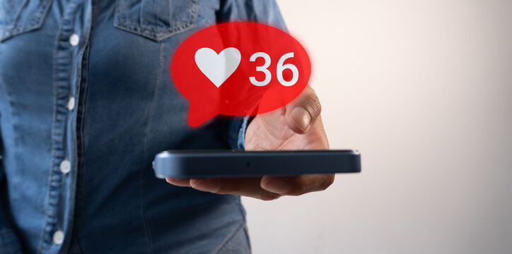 Close-up of a man's hand holding a smartphone with a red "like" or heart icon on a social media profile page and the white number "36" - Powered by Adobe