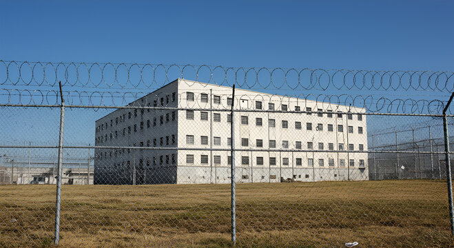 A large, imposing prison building is seen behind a barbed-wire fence under a clear blue sky.