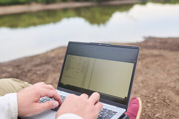 Unrecognizable person sitting outdoors on the bank of a stream, working on his laptop. Concept:...