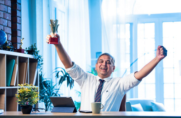 Mid age Indian businessman proudly holding trophy and smiling in stylish office