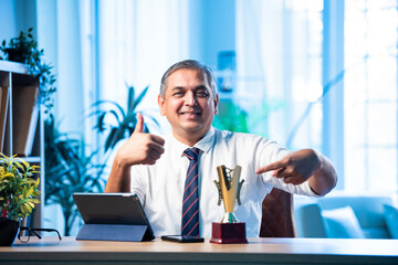 Mid age Indian businessman proudly holding trophy and smiling in stylish office