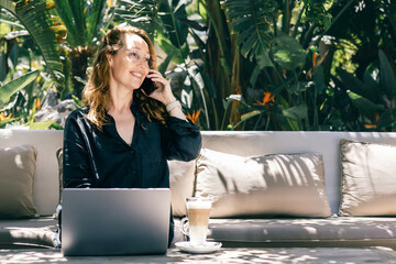 Smiling Woman Talking on Phone While Working on Laptop Outdoors in Tropical Garden with Coffee