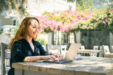Smiling woman working remotely on laptop in sunny outdoor cafe with blooming flowers in background