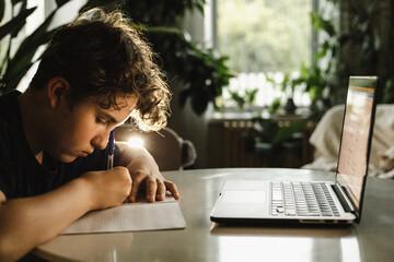 Teen boy writing in notebook with pen, studying at table with laptop, home interior daylight
